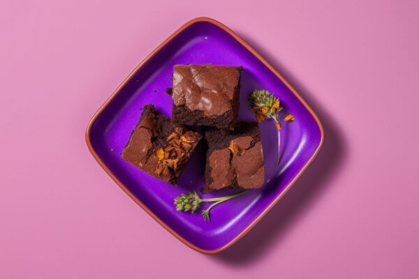 Brownies next to marijuana buds on a purple dish on a pink background.
