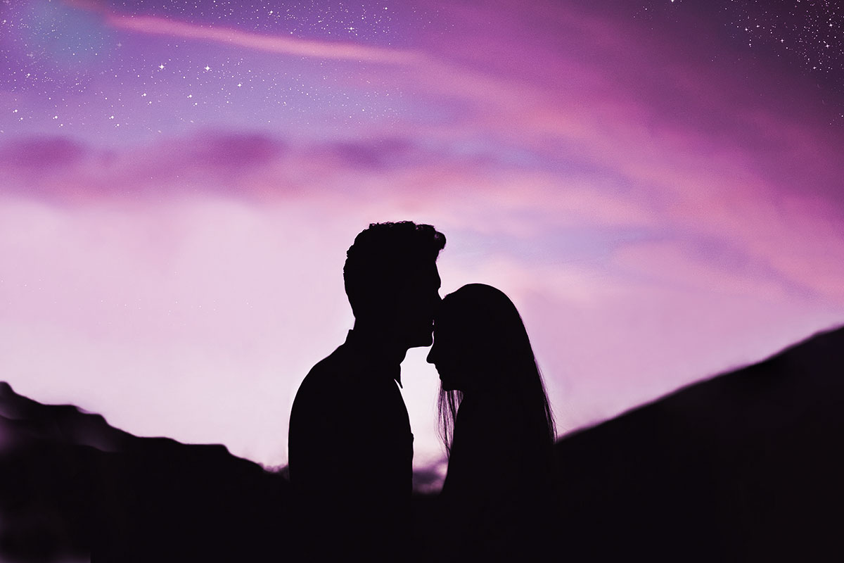 Silhouette or a man kissing a woman's forehead on a purple afternoon background.