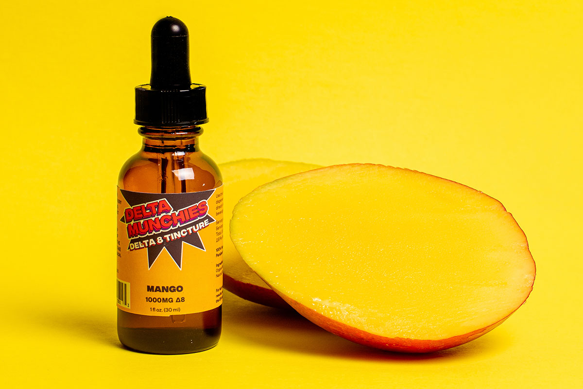 Bottle of Delta Munchies' Mango delta 8 THC tincture next to a slice of mango on a yellow background.