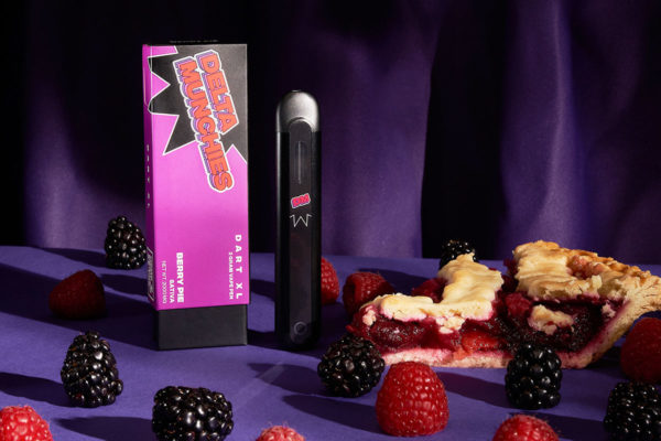 Delta Munchies' Berry Pie delta 8 disposable standing on a purple next to mixed berries and a slice porfavor berry pie.