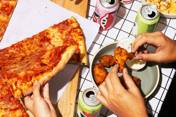 Women eating cheese pizza and chicken wings with canned drinks on a checkered table.