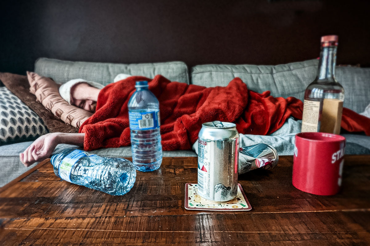 Man lying on a couch covered with a red blanket behind a table with water bottles, beer can, and an alcohol bottle.