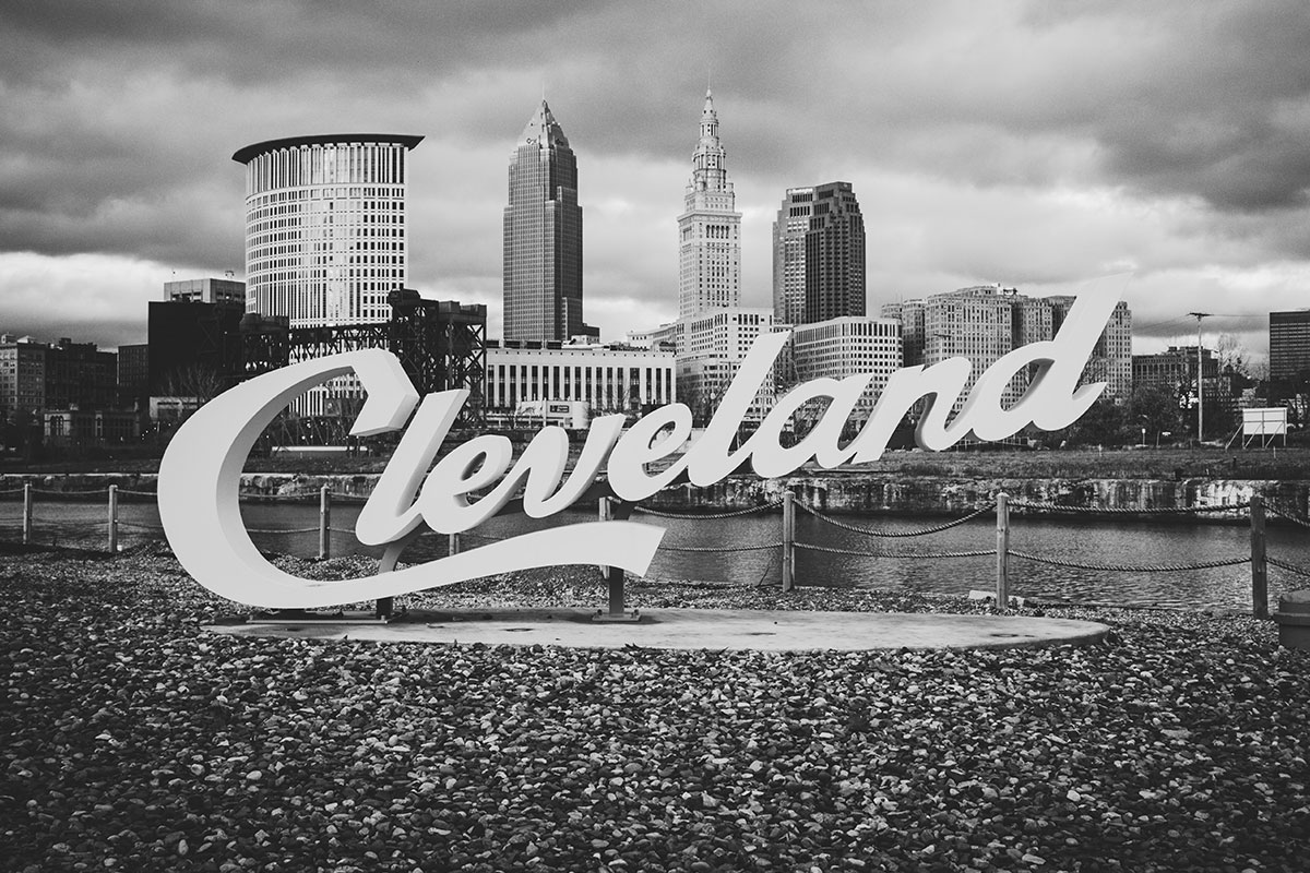 Black and white photo of a sign that says "Cleveland" with downtown Cleveland behind it.
