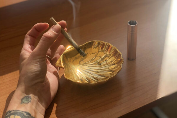 Man's hand holding a lit infused pre roll over a seashell shaped ashtray.
