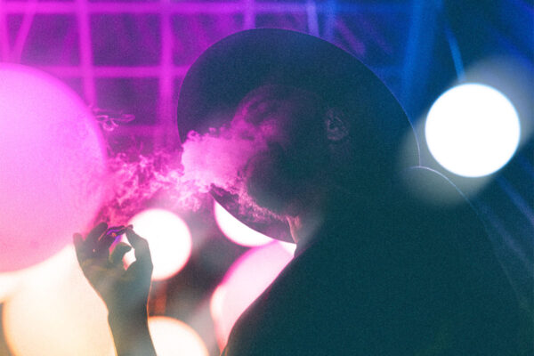 A man wearing a hat smoking a lit joint standing at a place with blue and pink neon lights