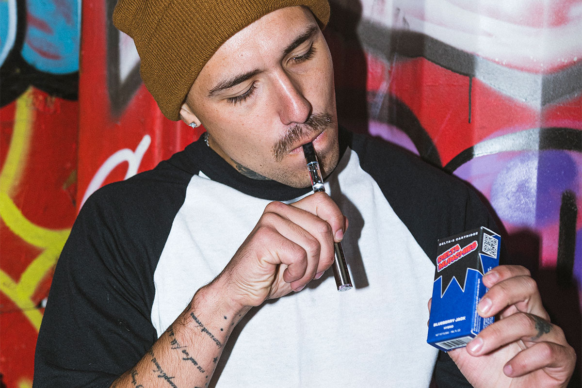 A man holding a pack of Delta Munchies cartridge in his left hand and holding a cartridge in his right hand up to his mouth