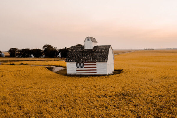 A white-colored tiny house on a farm field with a US flag on its outer wall and trees in the back