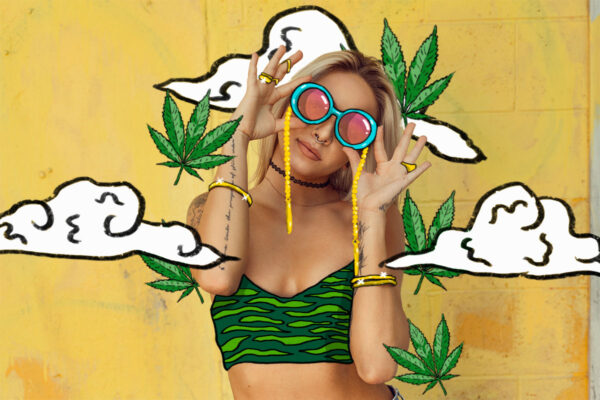 A woman holds goggles over her eyes amidst marijuana leaves on a vibrant yellow background