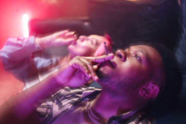 A man and a woman, smoking a weed together in a dimly lit environment with a reddish hue