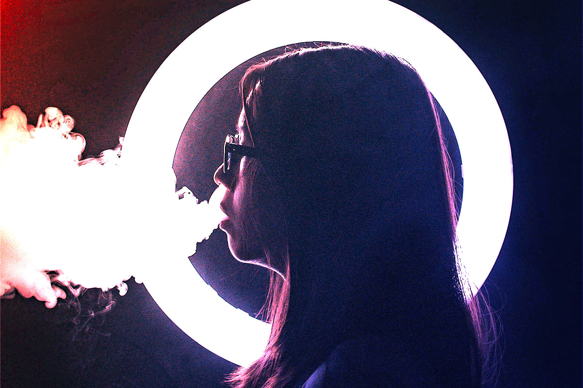 A girl wearing glasses exhales smoke in front of a bright ring light, set against a dark background