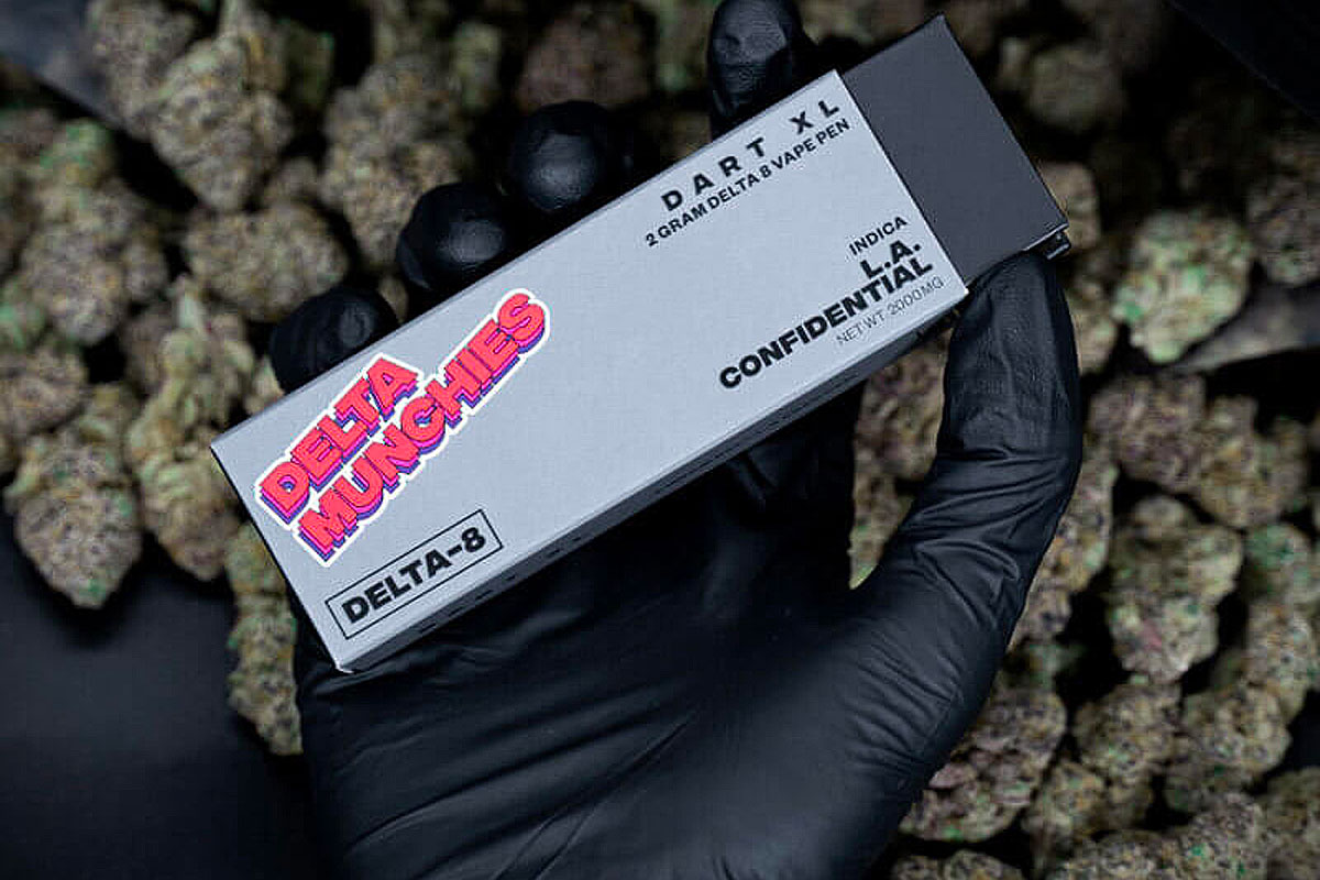 Black-gloved hand holding a pack of Delta Munchies Dart XL 2g Delta 8 vape pen - Indica L.A. Confidential, with cannabis plants in the background