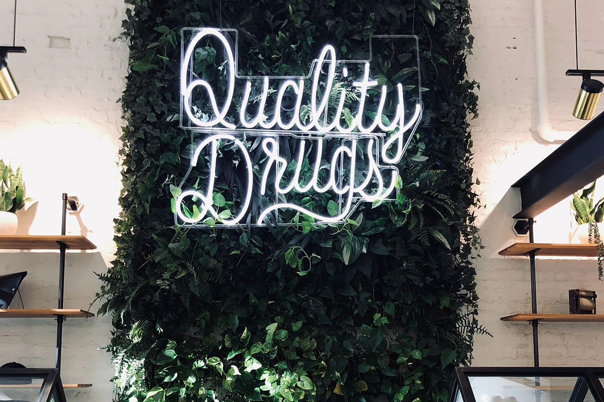 A neon 'Quality Drugs' sign contrasts against a backdrop of fresh green leaves on a clean white wall