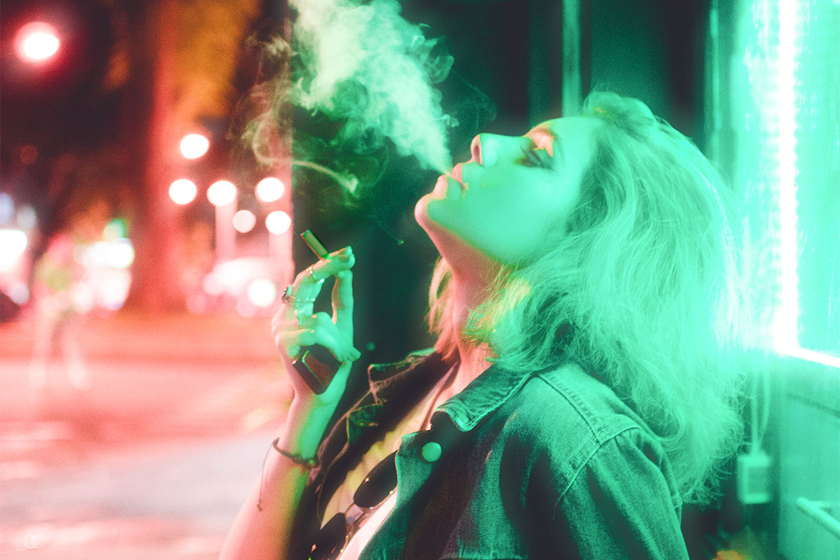A woman smoking a joint on the street, wearing a jacket and sunglasses hanging on her T-shirt with a greenish light illuminating her face and body