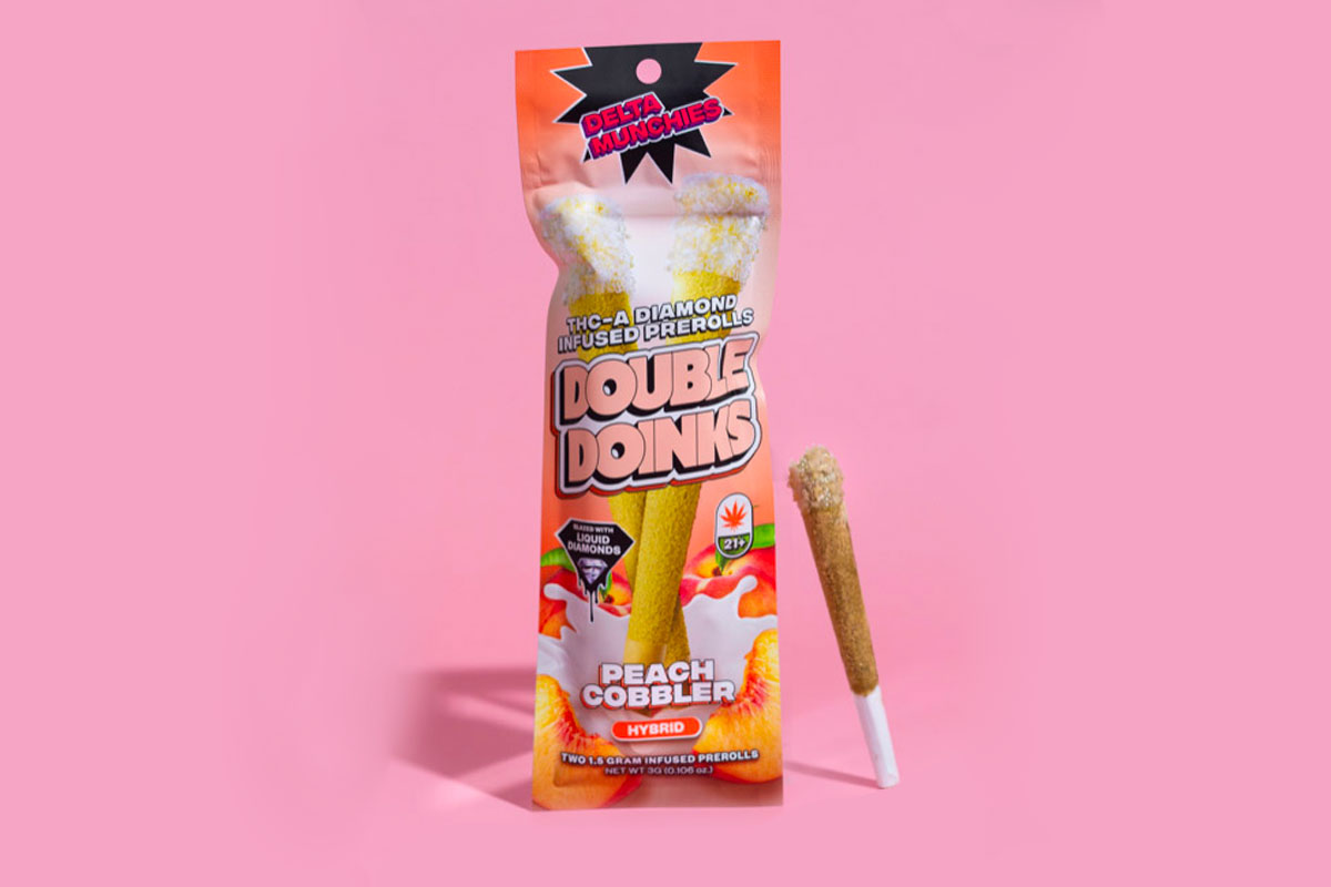 On a pink background, there is a pack of THC-A diamond-infused prerolls called Double Doikins, which have a peach cobbler flavor, with a single preroll placed beside the pack