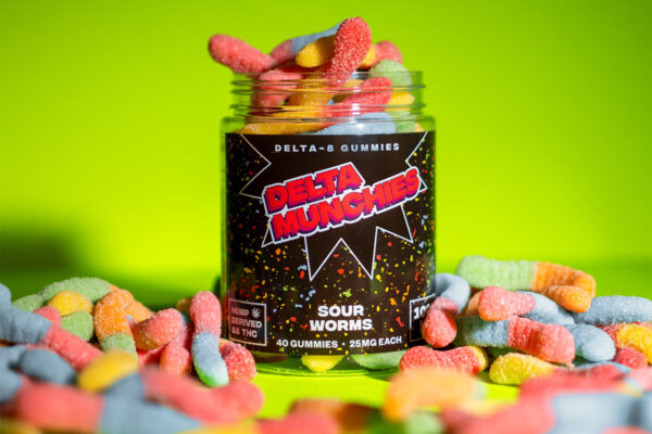 A container of 25mg Delta Munchies Delta-8 gummies with Sour Worms flavor, surrounded by more gummies on a green surface