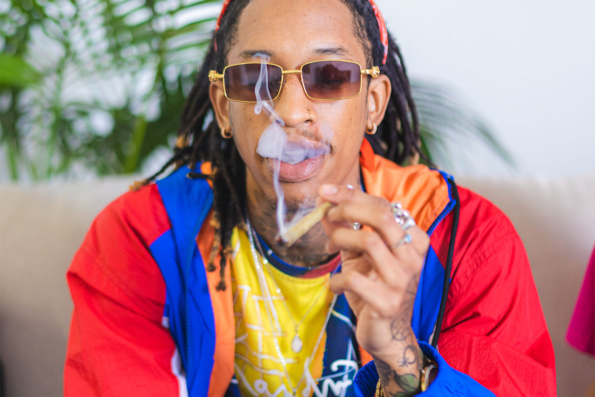 Long-haired man with tattoos, sunglasses, holding a joint, exhaling smoke, with palm tree leaves in the background