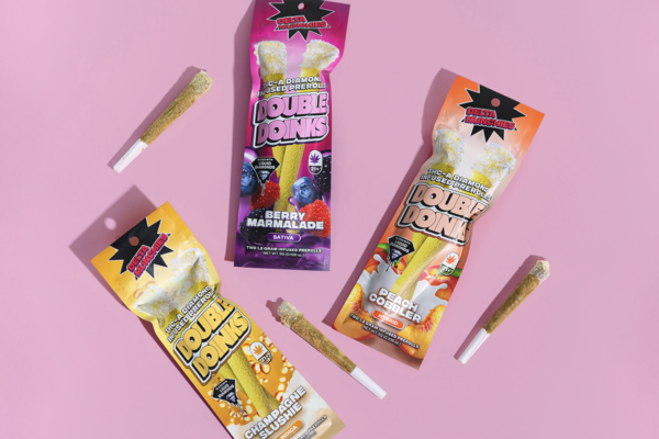 Three Delta Munchies Double Doinks prerolls with Champagne Slushie, Berry Marmalade, and Peach Cobbler flavors on pink surface, with three additional prerolls beside them