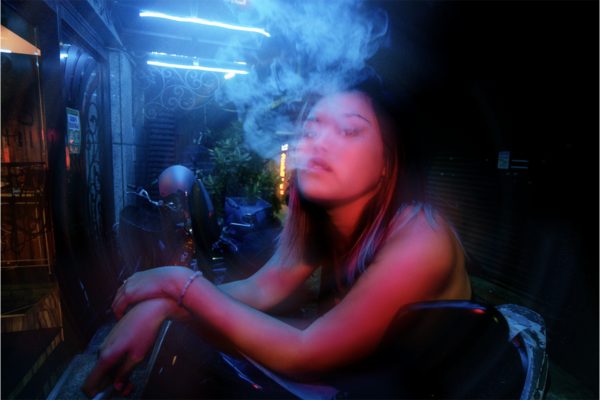 A girl sits, with a pink light falling on her face, bluish light illuminating her backside, while she exhales smoke