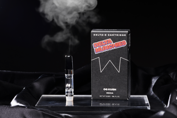 Delta Munchies Delta-8 cartridge OG Kush Indica next to a cartridge, exhaling smoke, on a sleek black glass surface