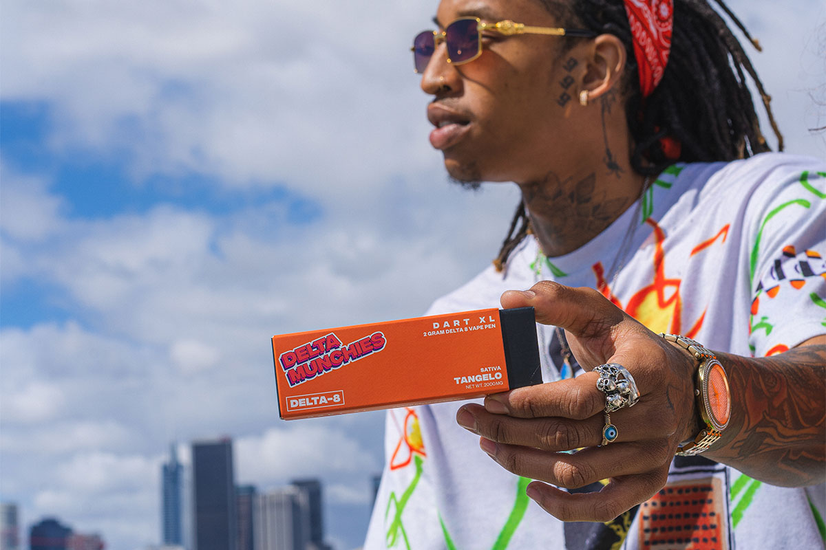 A man, sporting sunglasses, holds a package of the Delta Munchies Dart XL Delta 8 vape pen in Sativa Tangelo flavor, set against a backdrop of buildings and a blue sky.
