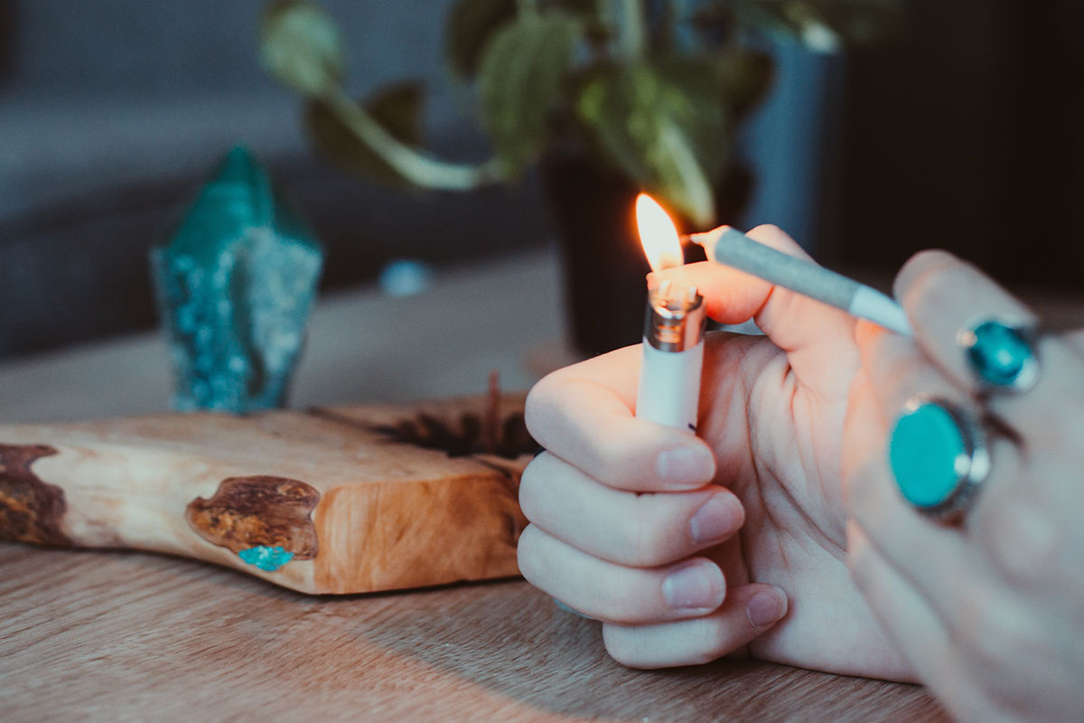 A hand is holding a cannabis joint and a lighter in the other hand, with a backdrop of trees and woods behind