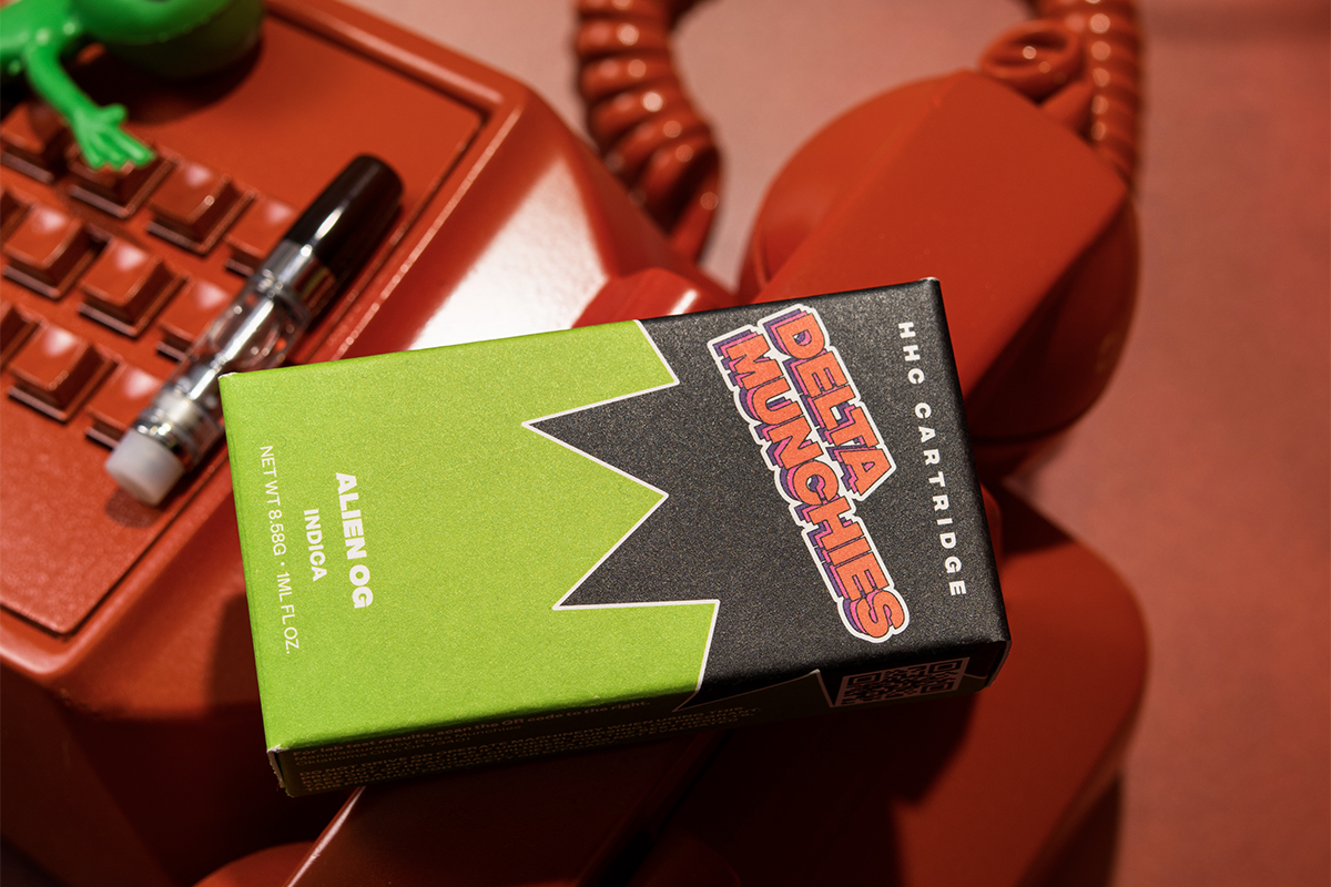 A package of Delta Munchies HHC Cartridge in Alien Og Indica flavor is placed on a red telephone, accompanied by a cartridge